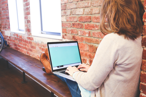 business woman on her computer on a bench leaning against a wall with her feet on the bench