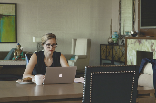 woman on her apple computer, sitting at a kitchen table, with a mug and a muffin